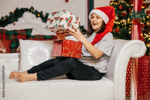 Excited woman opening Christmas gifts while wearing a Santa hat in a festive living room