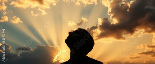Silhouette of a man looking up at a dramatic sky with sun rays. Hope, faith, and spirituality concept. Backlit person against a golden sunset background