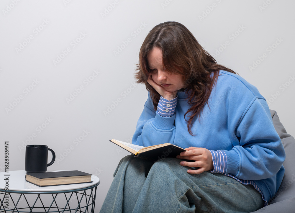 Obraz premium Young woman sitting indoors and reading a book with deep focus, leaning her chin on her hand while