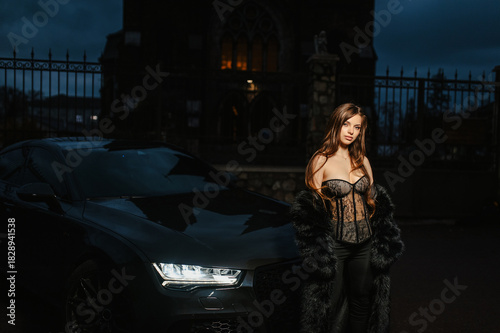A photograph, a portrait of a beautiful young sexy brunette woman, a stylish model in lingerie, in a fur coat, standing at night in the dark with a chic expensive black car on a city street.