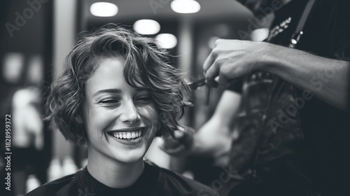 A woman with curly hair smiles brightly while sitting in a salon chair. A hairstylist carefully cuts her hair and soft lighting creates a warm atmosphere.