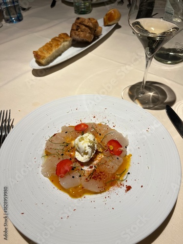 Gourmet White Fish Carpaccio or Ceviche Served on a Minimalist Plate with Citrus Dressing, Edible Flowers, Spices, and a Glass of White Wine in Fine Dining Setting