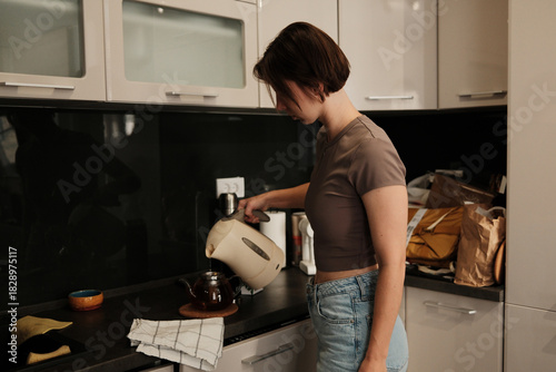 A woman pours hot water from an electric kettle into a glass teapot, preparing a warm drink in a calm kitchen setting.