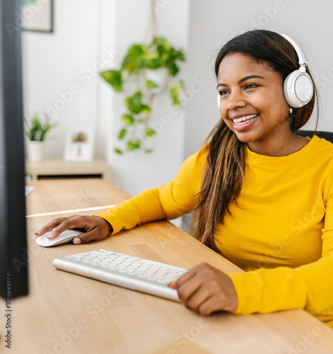 Happy young woman working from home, enjoying music with wireless headphones while using a desktop computer