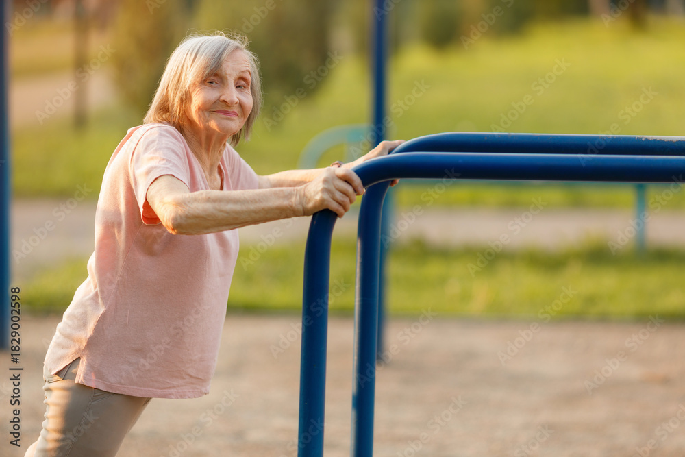 Obraz premium A happy 87-year-old senior woman performing an assisted push-up or leaning exercise on blue outdoor parallel bars, looking at the camera. Healthy and strong retirement.