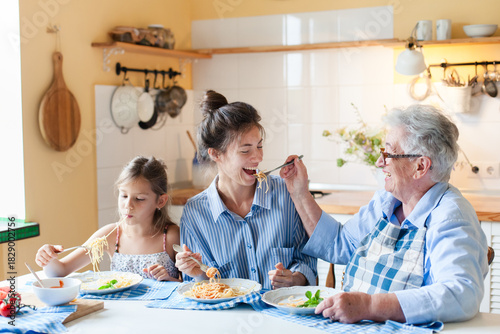 Happy multigenerational family eating pasta together at home kitchen, having fun. Grandmother feeding adult daughter, child enjoying comfort food. Candid, authentic lifestyle, joyful mealtime moment