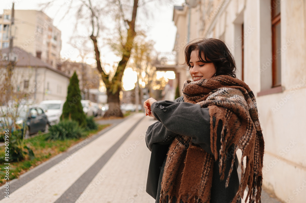 Fototapeta premium smiling woman holding gray cat on sidewalk, wrapped in layered shawl and overcoat, strolling past boutique