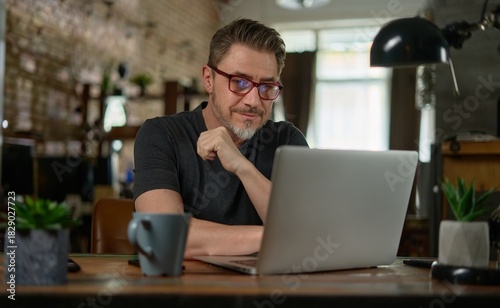 Bearded man working online with laptop computer at home sitting at desk. Home office, browsing internet. Portrait of mature age, middle age, entrepreneur.