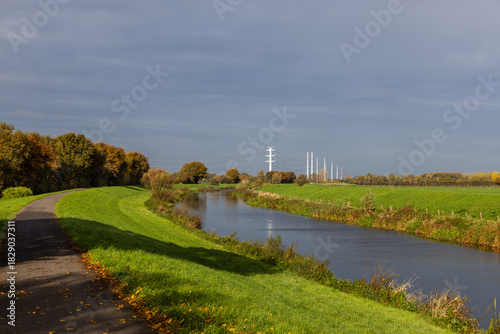Winding paved path beside calm river, autumn trees and green fields under dramatic cloudy sky, power lines stretch across serene rural landscape blending nature and infrastructure.