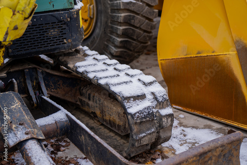 Detailed winter scene showing a compact excavator rubber track with snow buildup alongside a large yellow wheel and construction bucket on frozen ground