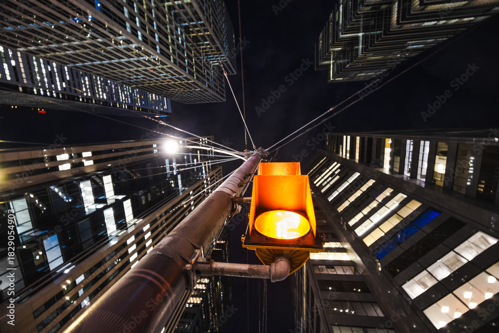 Fototapeta premium A glowing urban traffic light hangs above the street like a small sun, framed by looming skyscrapers that tilt inward, creating a dramatic tunnel of glass and steel against the night sky.