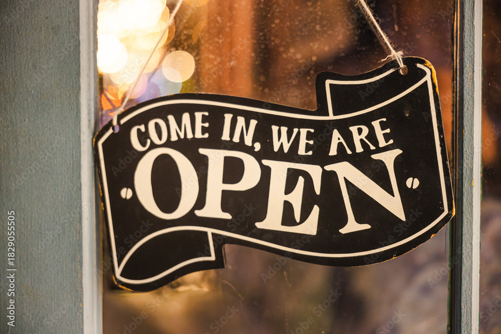 Fototapeta premium Closeup of a vintage open sign hanging on a shop window with warm bokeh lights in the background.