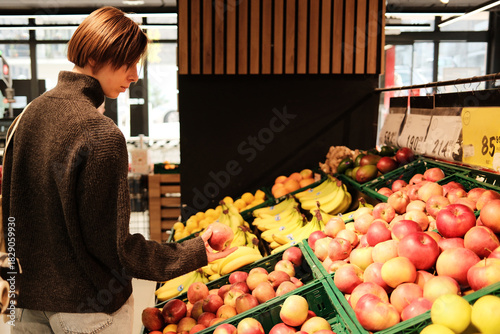 A woman holds an apple while standing at the fruit display in a grocery store. The concept shows everyday shopping and healthy food selection.