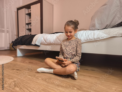Young girl sitting on wooden floor in cozy bedroom, smiling while using smartphone, surrounded by soft bedding and warm lighting, showcasing modern lifestyle and technology