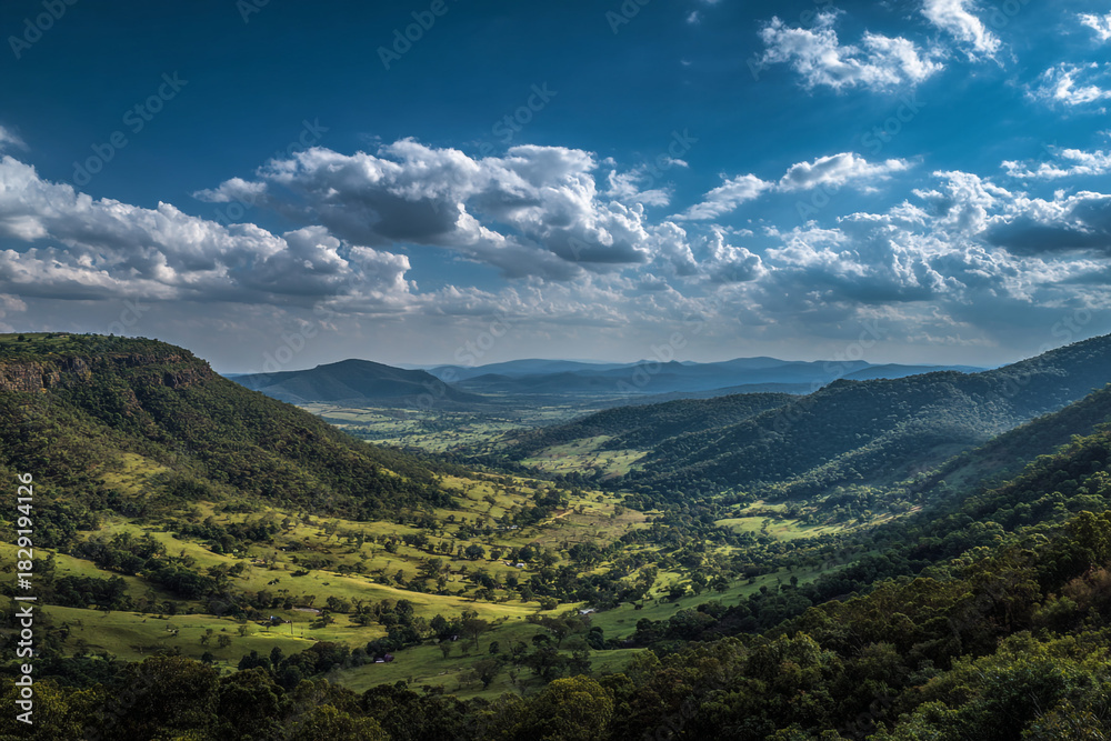 Fototapeta premium Lush green rolling hills stretch towards distant blue mountains under a dramatic sky filled with white clouds