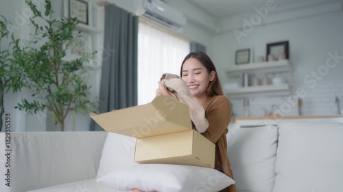 Happy young Asian woman opening delivery package at home. Beautiful female customer open box and sitting on couch in living room