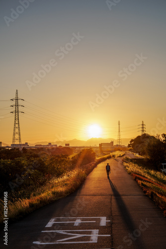 Foto Setting sun touches distant mountain top, casting long shadows across expansive landscape featuring power towers and tall grass embankments enveloping riverside path with locals enjoying gold evening