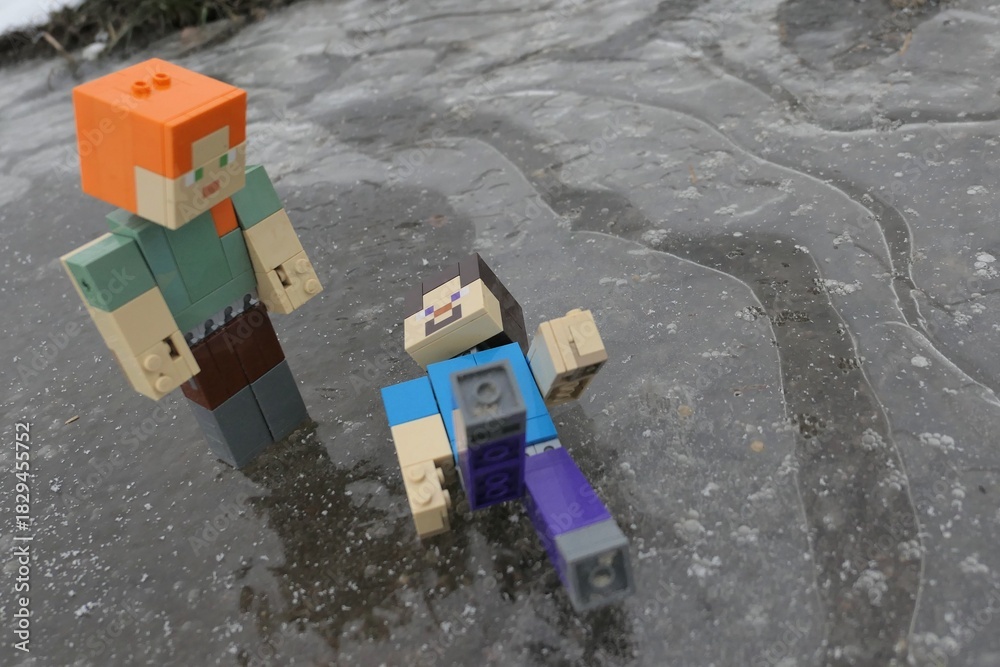 Naklejka premium LEGO Minecraft action figure of Steve accidentaly fallen on icy surface while gliding with Alex, standing above him. Air bubbles visible under frozen ice puddle.
