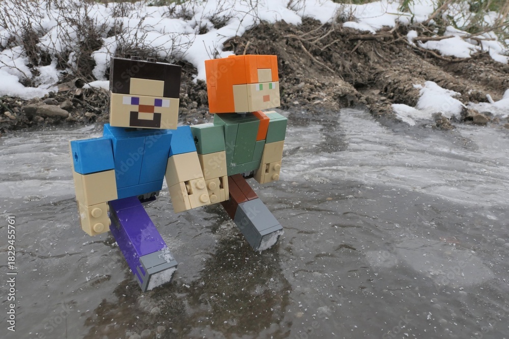 Naklejka premium LEGO Minecraft action figures of main characters Steve and Alex gliding on ice of frozen puddle on edge of agricultural field, snowy puddle bank and frozen mud in background.