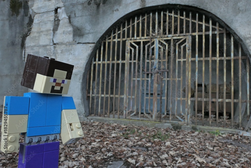 Obraz premium LEGO Minecraft large figure of main character Steve standing in front of rusty gate to a mine or mining storage in village Hodrusa - Hamre, central Slovakia, opposite abandoned Sandrik factory