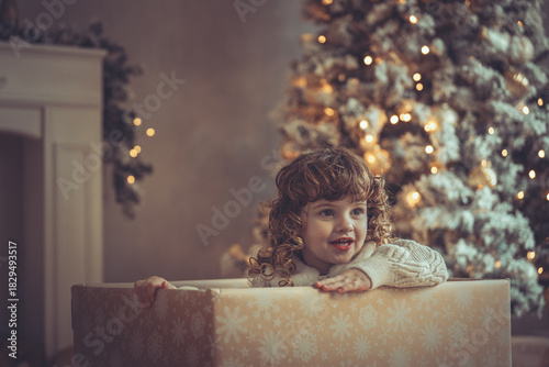 Hübsches kleines Mädchen mit Locken sitzt in einem Geschenkkarton vor dem Weihnachtsbaum zu Weihnachten, Abendstimmung mit Lichtern Var. 1