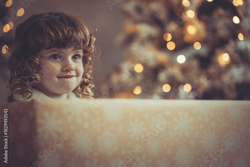 Hübsches kleines Mädchen mit Locken sitzt in einem Geschenkkarton vor dem Weihnachtsbaum zu Weihnachten, Abendstimmung mit Lichtern Var. 2