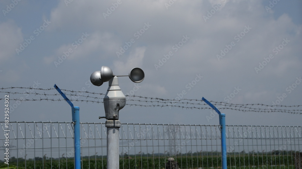 Obraz premium Anemometer measures wind speed at weather station near barbed wire fence on partly cloudy day, showcasing technology and environmental monitoring.