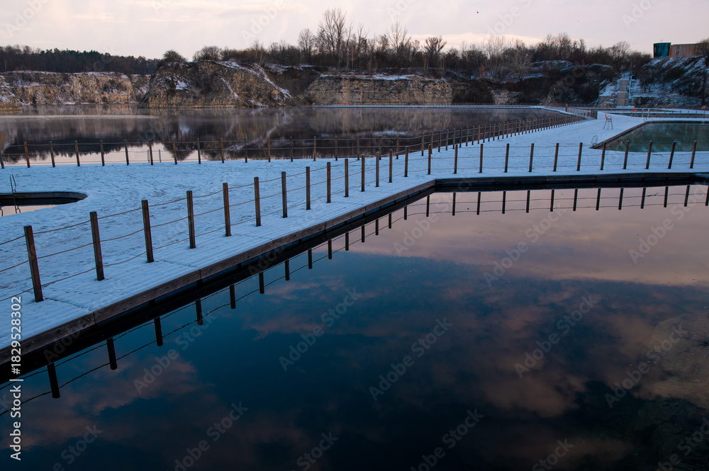 Naklejka premium Snow-covered walkway over a calm lake with reflections of clouds.
