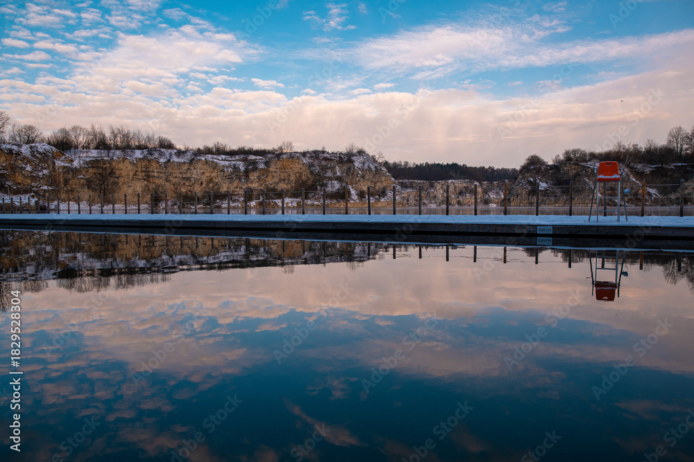 Fototapeta premium A tranquil outdoor swimming pool reflecting a cloudy sky and surrounding trees.