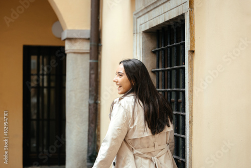 Young woman with long dark hair wearing a stylish trench coat, smiling while walking through a charming urban alleyway, showcasing a vibrant and inviting atmosphere Free Download Young woman with long dark hair wearing a stylish trench coat, smiling while walking through a charming urban alleyway, showcasing a vibrant and inviting atmosphere - GFXLoad