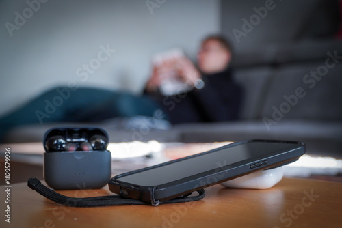 Smartphone and headphones set aside in the foreground while a man reads a book in the background. Concept of digital detox, disconnecting from technology, and returning to analog leisure