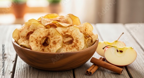 A wooden bowl filled with crispy, golden apple chips, garnished with cinnamon sticks and fresh apple slices on a rustic wooden table.