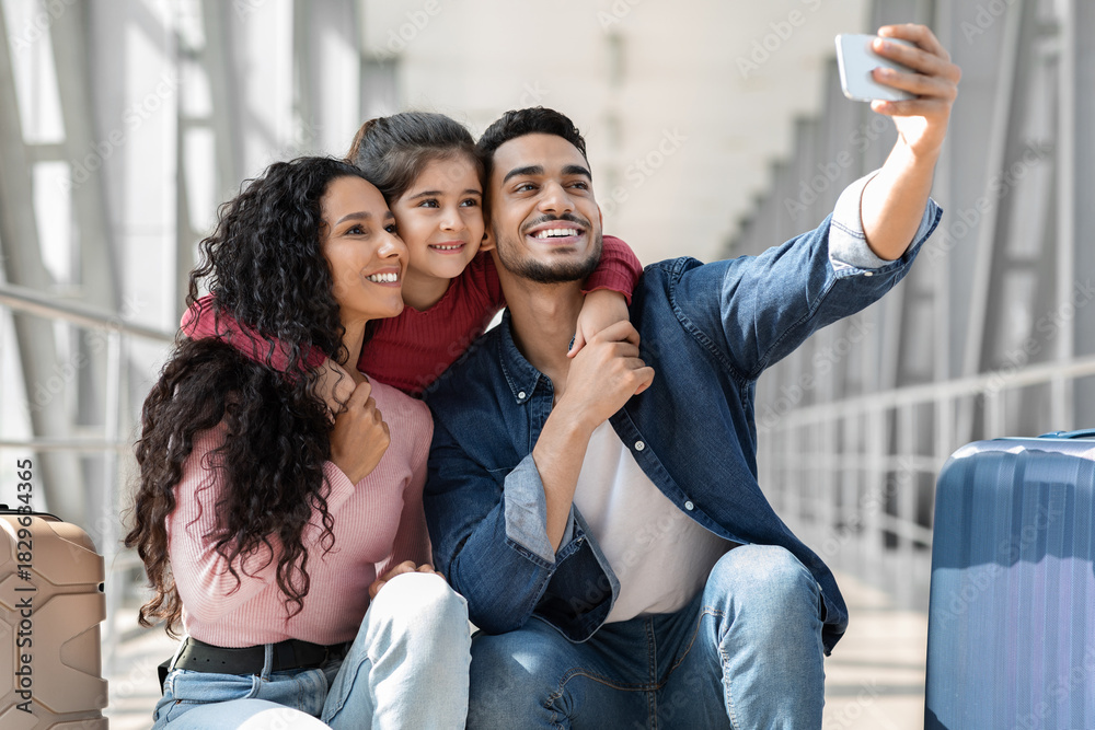 Obraz premium A happy family sits together at an airport terminal. They are smiling and posing for a selfie, showcasing their close bond. Luggage is placed near them, indicating travel plans.