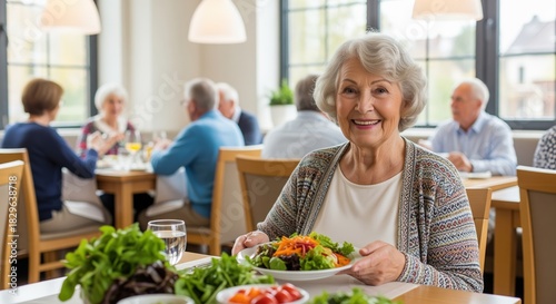 A smiling elderly woman enjoys a healthy meal with fresh vegetables in a bright dining room. Other seniors socialize in the background. Ideal for retirement and senior lifestyle content.