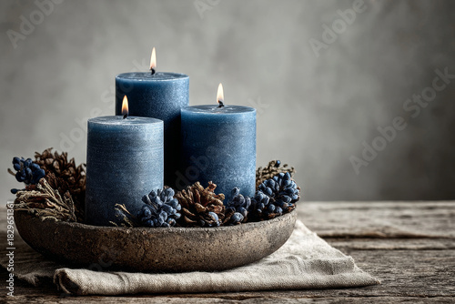Three dark blue wax candles burning in rustic bowl with dry pine cone decoration on wood table for cozy winter home atmosphere