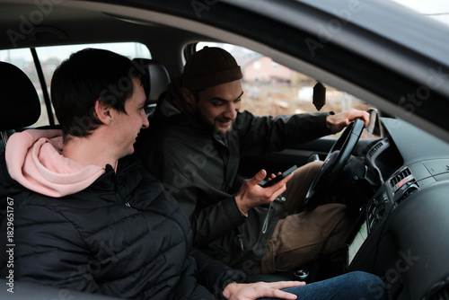 Two men sit inside a car while one checks information on a smartphone. The scene reflects navigation, planning, and road assistance.
