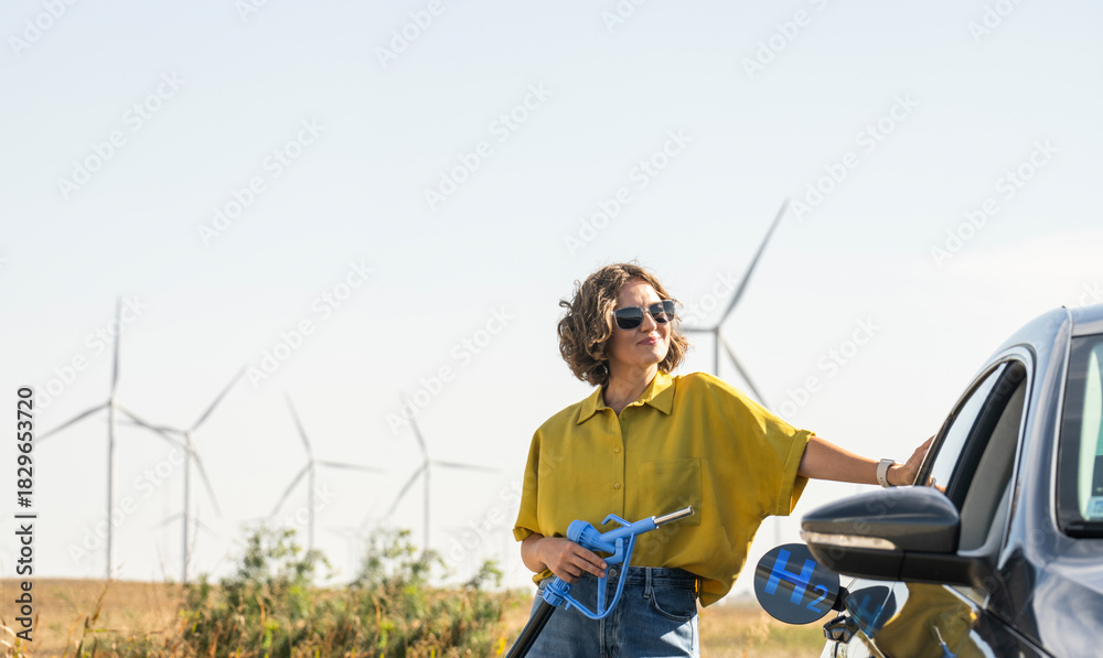 Naklejka premium Woman holds a hydrogen fueling nozzle. Refueling car with hydrogen fuel. Wind turbines in the background