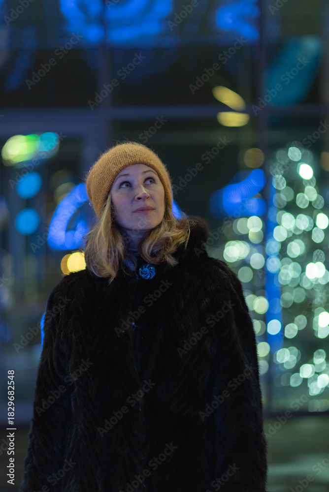 Fototapeta premium Woman with long blonde hair, dressed in a black fur coat, stands outdoors surrounded by colorful holiday lights, creating a festive atmosphere during winter season
