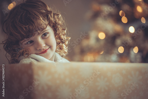 Hübsches kleines Mädchen mit Locken sitzt in einem Geschenkkarton vor dem Weihnachtsbaum zu Weihnachten, Abendstimmung mit Lichtern Var. 9