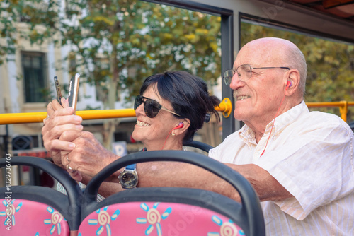 Smiling senior couple of tourists enjoying a sightseeing tour on an open-top bus sitting together listening to audio guides through earphones taking photos. A moment of adventure in relaxed atmosphere
