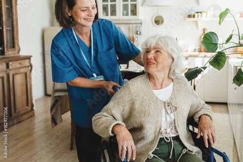 Smiling female caregiver pushing senior lady in wheelchair at retirement home or geriatric clinic after having lunch in kitchen, friendly assisting old disabled woman. Social service concept