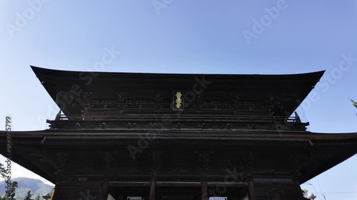A tall, Japanese-style wooden gate under a blue sky, the entrance to Zenkoji Temple in Nagano, Japan.