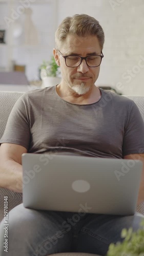 Older man working online with laptop computer at home sitting on couch in living room. Home office, browsing internet. Portrait of happy, mature age, middle age, mid adult man in 50s, smiling.