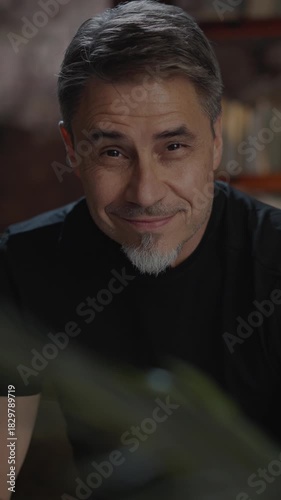 Confident older white man at home, sitting on couch in dark living room. Portrait of happy, mature age, middle age, mid adult caucasian man in 50s, smiling, looking at camera.