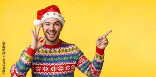 Cheerful man in a colorful Christmas sweater and Santa hat giving a thumbs up while standing in front of a yellow background