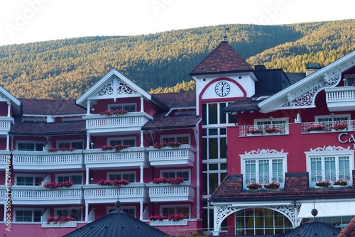 A stunning view of a pink hotel in Ortisei, Italy, with beautiful flower boxes on the balconies, a clock tower, and the green mountain backdrop under a clear sky.