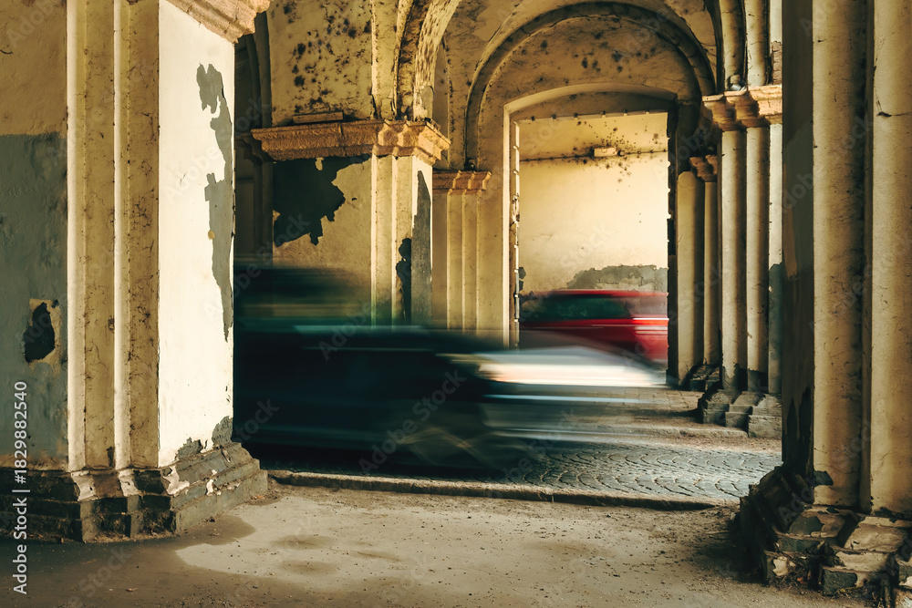 Fototapeta premium Motion-blurred vehicles speeding through a weathered arched passageway, contrasting modern traffic with aged, crumbling architecture.