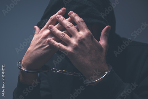 Low-key portrait of a hooded criminal with hands raised to an obscured face, handcuffs visible and dramatic shadows emphasizing tension and guilt.