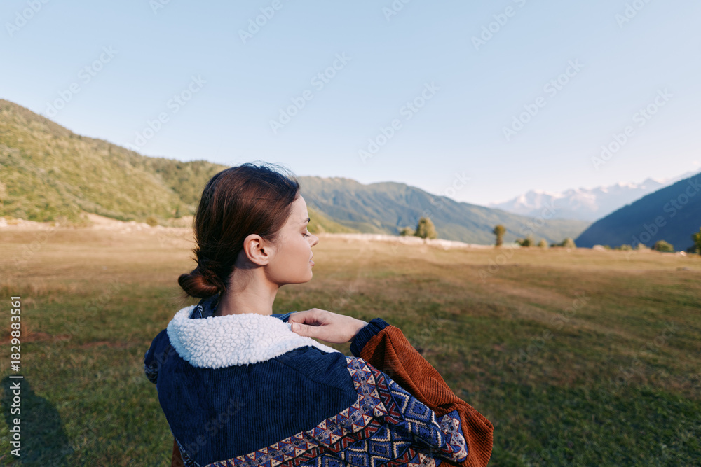 Naklejka premium Woman in a warm jacket with fleece collar stands in a wide meadow overlooking distant mountains, profile view outdoors. Travel portrait capturing landscape, calm horizon and soft autumn light.