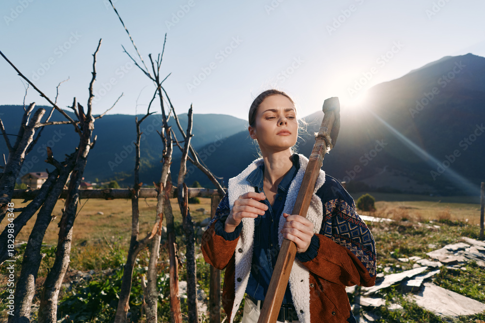 Fototapeta premium Woman farm tool gardening mountains sunlight in a rural outdoor portrait, holding a wooden pitchfork by a fence with calm expression during countryside agricultural work.